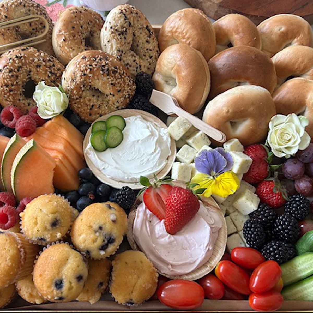 Assorted bagels, muffins, fruits, and vegetables on a platter with a small bowl of cream cheese.