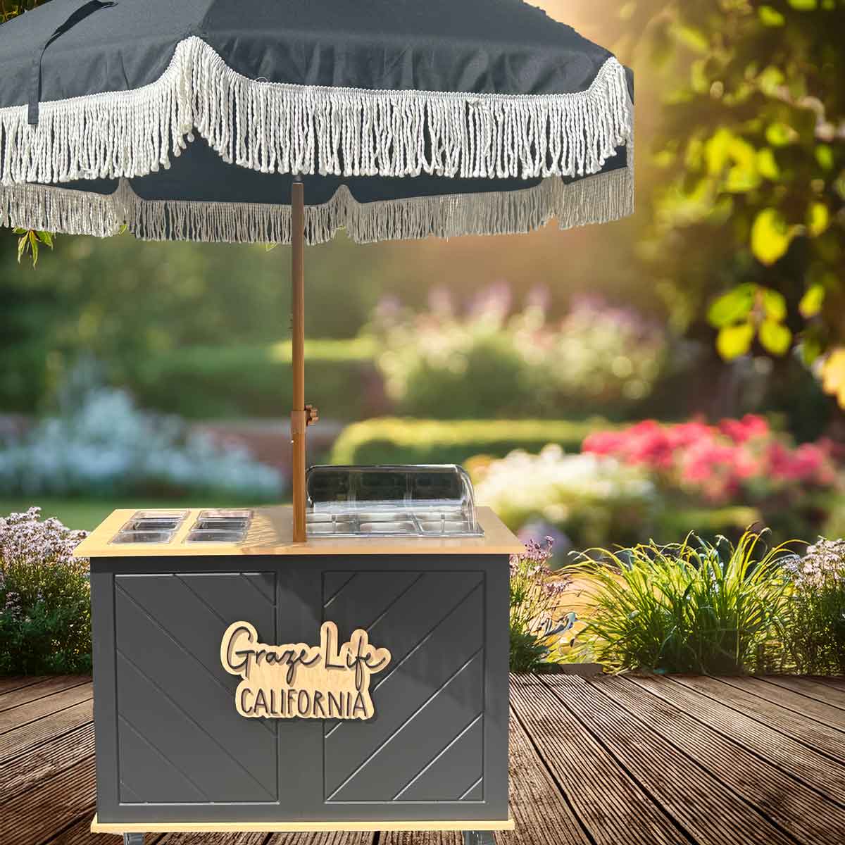 Charcuterie Cart with 'Graze Life California' branding under a black umbrella with white fringe in a garden setting.