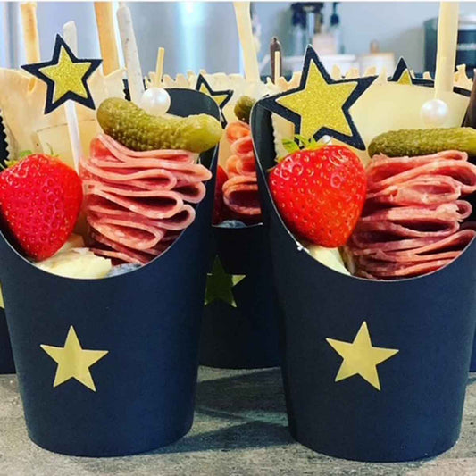 Two navy blue Charcuterie cups with star decorations, filled with snacks including strawberries, salami, and pickles.