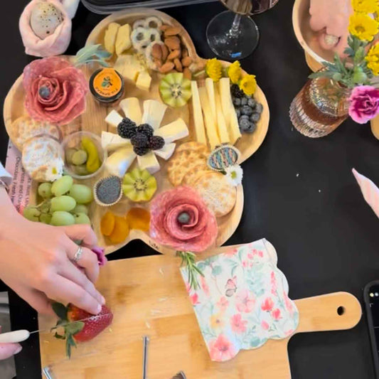 Wooden charcuterie board with fruits, cheese, and flowers on a black surface.
